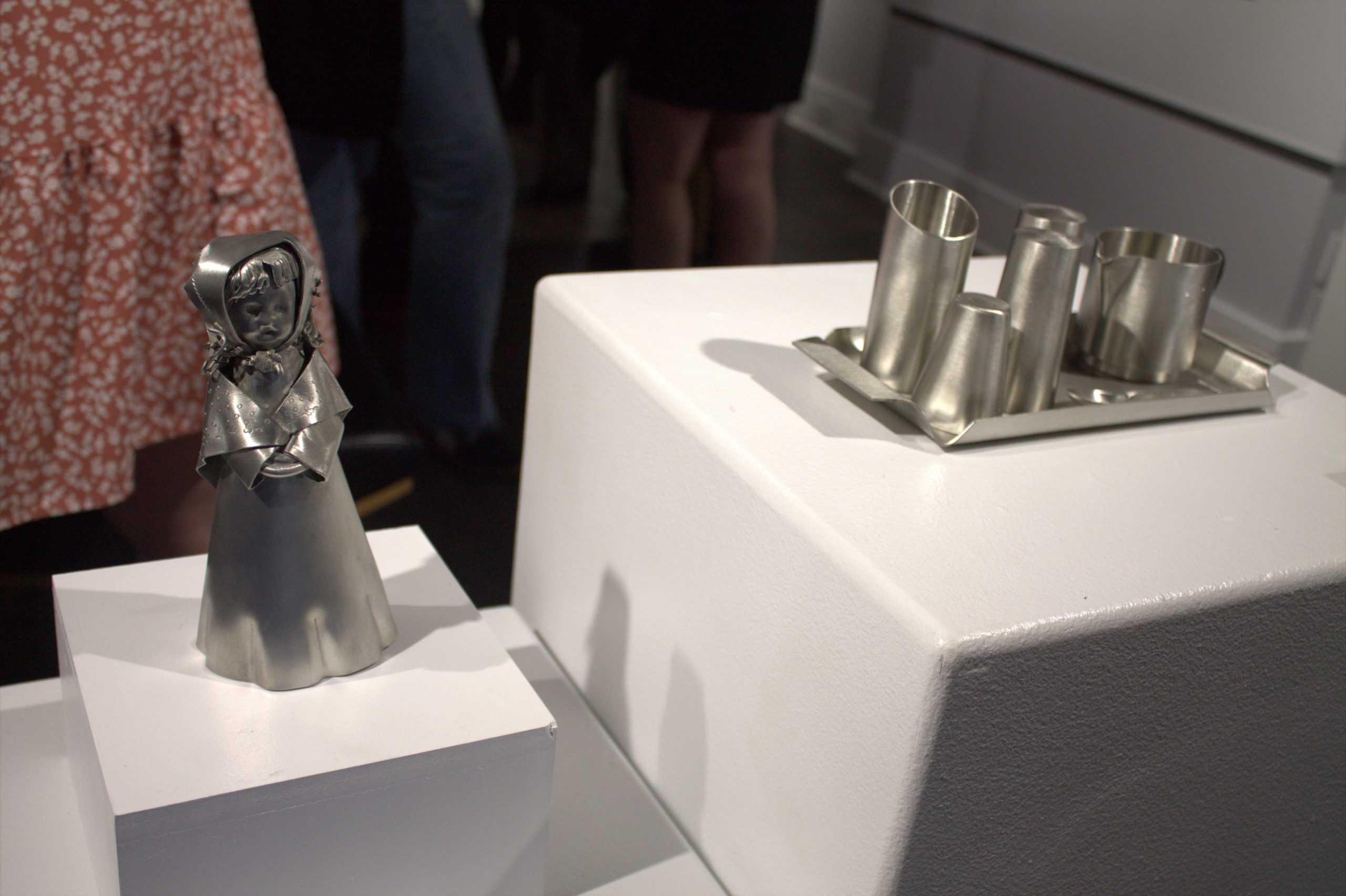Two works of pewter on display at an art gallery. One of a little girl and the other a collection of various cups.